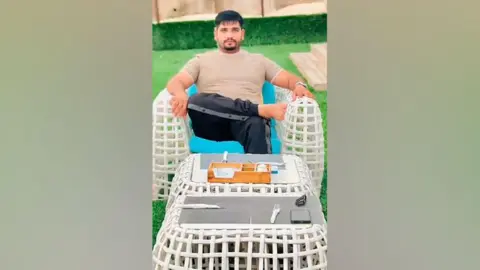 TikTok/Fadi Gujjar An image showing Fadi Gujjar at a beachside cafe. He is wearing a dark tracksuit and a t-shirt and is sitting on a white chair. In the foreground the table is set for dinner.