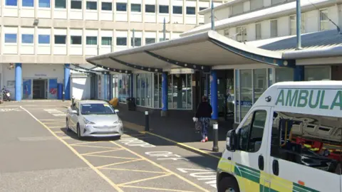 Google The outside of North Tees Hospital An ambulance is waiting in the yellow hatched drop off zone and a silver car is also parked. Parts of larger hospital building can be seen in the background.