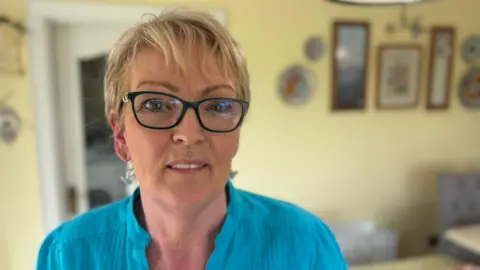 A woman stands in her kitchen. She has blonde hair, glasses and is wearing a blue shirt.