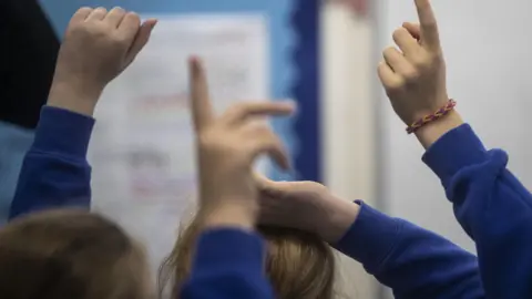 Getty Images Photo of hands going up to ask a question in a primary school class. Four hands are visible.
