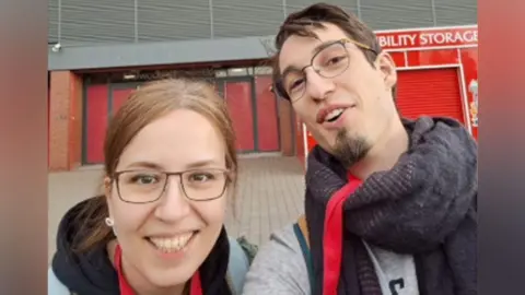 The University of Liverpool Dr Farnaz Kamranzad (L) has brown hair pulled and is wearing glasses and a black hooded top. Dr Antoine Septier (R) has short brown hair and is wearing glasses and a scarf. BOth are stood outside Anfield football stadium and smiling at the camera.