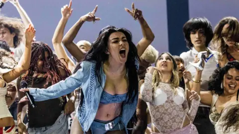 Getty Images Charli XCX performing on stage at the Grammy's. The singer wears a denim look made up of a bra top and pants under a long-sleeved matching jacket. She sings into the crowd, her long dark hair worn loose. She's surrounded by dancers.