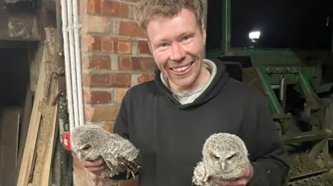 CPS Carruthers, with short blond hair and wearing a black hoody, smiles as he holds two young grey owls, one in each hand. Behind him is a wall and agricultural equipment