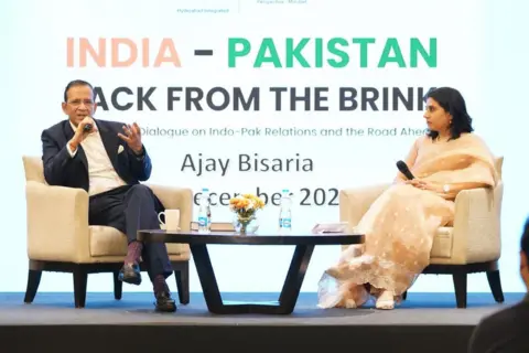 Ajay Bisaria Ajay Bisaria sits on the podium holding a mic in his hand.