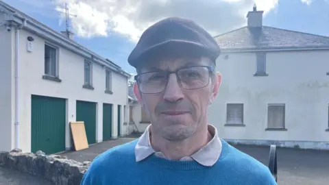 Sean Faloon standing in front of Clonduff parochial house on a sunny day in spring 2025. He has short, greying dark hair and is wearing a dark flat cap, glasses and a blue jumper over a polo shirt.
