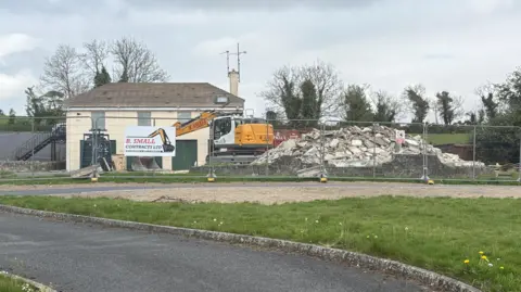 The former site of Clonduff parochial house. A large pile of rubble sits where the main building used to stand. A metal safety fence is blocking off the whole site. Roof tiles have been stripped from a smaller parish building in the background.
