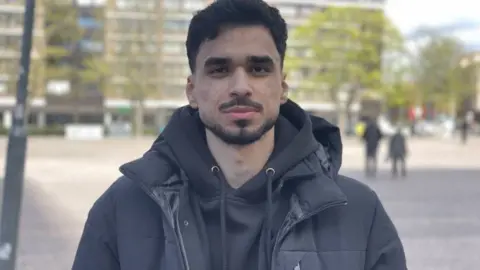 A man in a black hoodie and black coat looks at the camera. He is stood outside in a paved public area