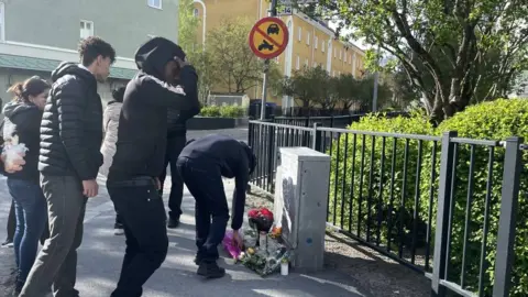 A group of people in dark clothing stand at a street corner. There is a pile of flowers on the ground and one person is laying more flowers down.