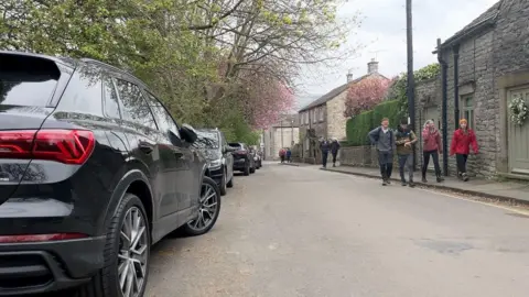 Cars parked in a street in Castleton with a group of hikers walking on a pavement opposite