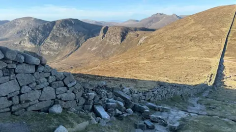 Dave Somerville A granite stone wall slopes down the side of mountain. A section of the wall has collapsed and a large number of stones are lying in rubble.