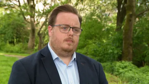 Thomas Kerr has swept back ginger hair, black-rimmed spectacles, a blue blazer and a light blue shirt. He is standing in front of grass and trees