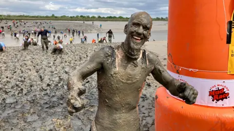 Chris Radburn/BBC Man covered from head-to-toe in mud. Lots of people behind him can be seen making their way across a very muddy river estuary.