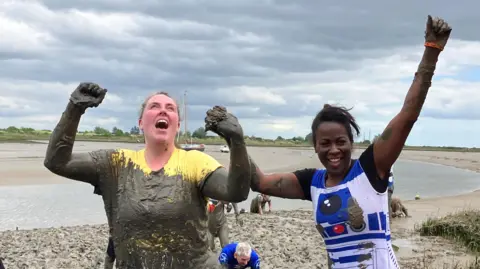 Chris Radburn/BBC BBC Essex presenters, Jodie Halford and Akylah Rodriguez cheer after making it through the mud. Jodie is particularly splattered.
