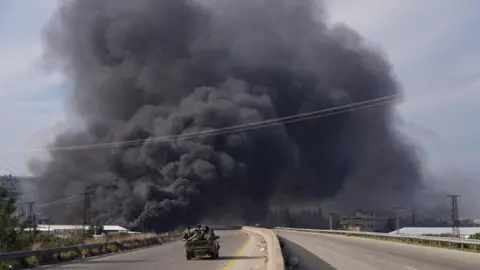 Reuters Smoke rises while members of the Syrian security forces ride on a pick-up truck during fighting with insurgents loyal to former president Bashar al-Assad, in Latakia (7 March 2025)