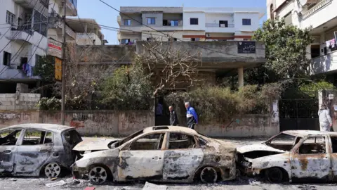 Reuters File photo showing people walking past burned-out cars in Jableh following the mass killing of Alawites in Syria's north-west coastal region (12 March 2025)