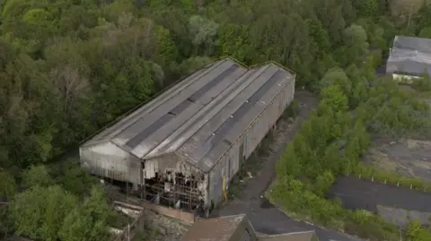 An aerial shot of the building where the fire took place. It is a long pair of buildings, somewhat like might be found in a railway yard, with triangular roofs and much of the cladding torn away at the near end. The front is severely damaged and it is surrounded by trees and other plant growth.