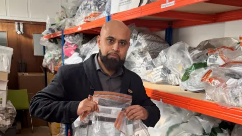 Trading Standards, London Borough of Barking and Dagenham Nazir Ali holds a clear plastic evidence bag up for the camera. He is standing in a room with shelves behind him that are filled with similar evidence bags. Nazir is a bald man, with a thick black beard. He is wearing a black jacket and a grey shirt.