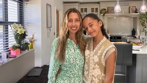 Chantelle Thompson, in a green floral dress, and Thailula Thompson in a white floral top, smile at the camera in the kitchen of their family home.