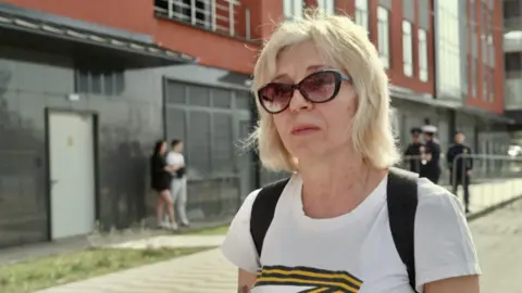 Blonde lady in her sixties wearing sunglasses stands in the street. The background is blurred but there are uniformed officers behind her.