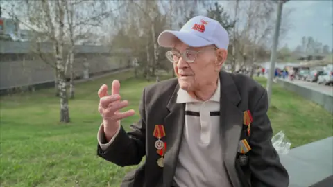 An old man wearing a cap with the 80th Anniversary of Russian victory logo. He is wearing a blazer covered in military medals.