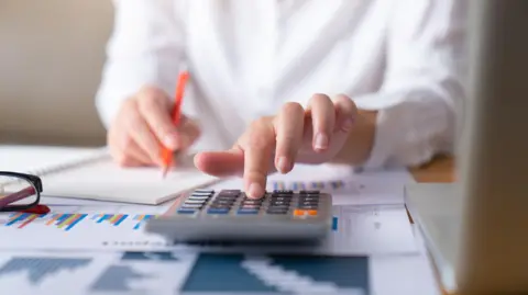 Getty Images Woman with spreadsheets and calculator in front of her. There is a laptop and glasses on the table. She is wearing a white shirt and holding an orange pencil while using the calculator.