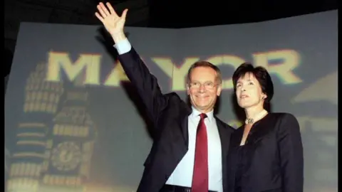 PA Media Jeffrey Archer and his wife Mary standing in front of a 'Mayor' backdrop. Jeffrey is wearing a dark suit and red tie and is waving. Mary is wearing a dark suit jacket and has short dark hair.