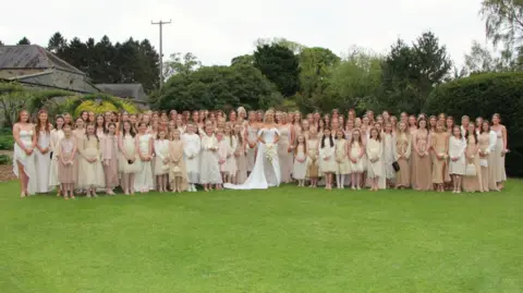 Mark Johnston A wide shot photo of a bride, with blonde hair, a white dress with an off the shoulder detail carrying a bouquet, stands in the middle of a large group of women and young girls in the front row, wearing a mixture of champagne, cream, white and tan dresses. The women are standing in the middle of a large garden, with tall trees and a leafy tunnel to the left and are looking at the camera.