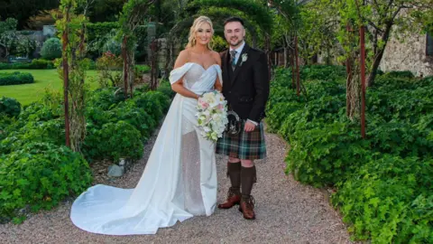 Mark Johnston The bride is wearing a white dress with off the shoulder sleeves and glitter details on the bodice and leg. She is standing on the left. The groom is wearing a kilt and suit jacket. They are standing beneath a garden arch and smiling at the camera.