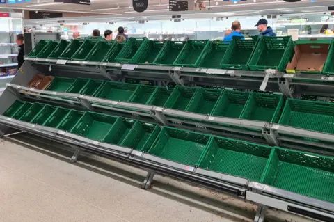 Radio Skye Empty green plastic boxes for fresh produce. Shoppers are browsing products in the background.