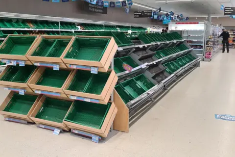 Radio Skye Empty green plastic boxes for fresh produce. There is a shopper browsing products in the background.