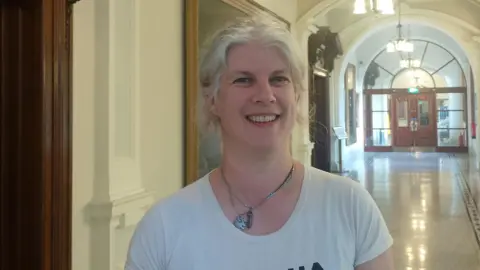 A woman, with white hair tied up, smiling at the camera. She is wearing a white t-shirt and a necklace. There is a long corridor behind her.