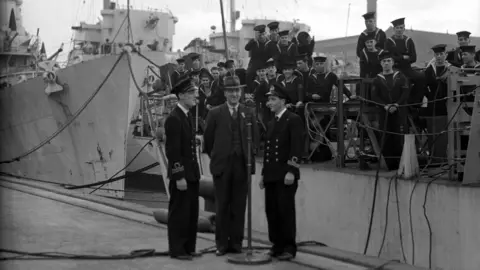 Commentator Lt. Commander Harry McMullan with Samuel Megraw, a shipyard worker and Lt. Commander J.M. Sowry, RNVR, broadcasting in Northern Ireland's contribution to Victory Report from Pollock Dock, Belfast