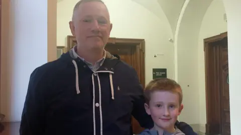 A man, with short brown hair, wearing a shirt and a navy jacket. He has his hand around a young boy, who has short red hair and is wearing a grey top.