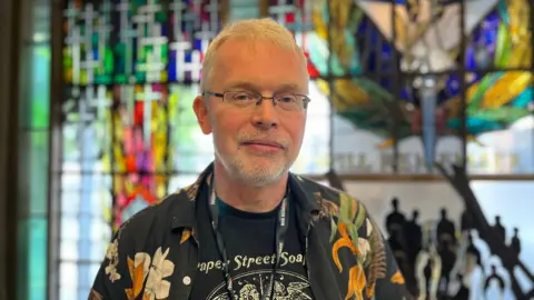 A grey haired man stares at the camera. He wears glasses and a hawaaian shirt.