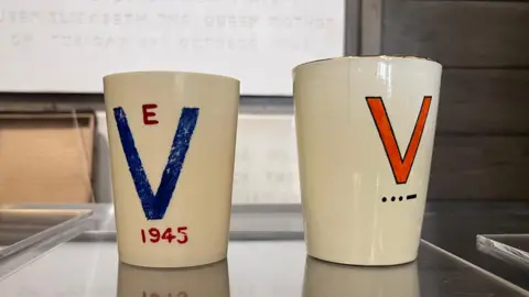 Two cups - both are white. The one on the left is painted with a blue V and the one of the right is painted with a red V.
