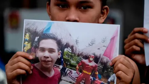 Getty Images A supporter of Kian Delos Santos attends a protest outside the police station where three policemen involved in Kian's killing were assigned in Manila on November 29, 2018. He is holding up a photo of a smiling Kian in a red shirt, standing near a superhero figurine.