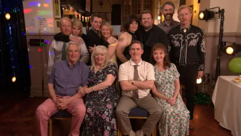 PA Images The main cast from Gavin and Stacey. Some of the cast members are sitting down on a chair and others are standing up. Everyone is smiling at the camera.
They are on the set of the final episode from last year