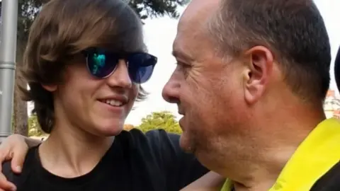 Michael Rainsford Mikey Rainsford, who has long brown hair and wears a pair of reflective sunglasses, poses for a picture with his father Michael Rainsford, who has thinning grey hair and is looking into his son's eyes.