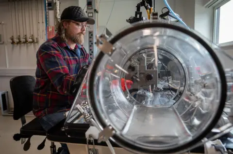 Tony Jolliffe/BBC News James Malley sits on a chair with his hands inside a long see-through tube. Inside are wires and metal pieces of equipment.