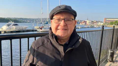 Liam is smiling at the camera and is wearing a grey flat cap. He has black framed glasses and is wearing a black water proof coat.