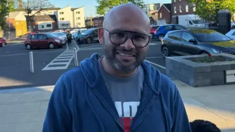 Blesson is msiling at the camera. He has black framed glasses and is wearing a navy zip up hoodie and grey t-shirt.