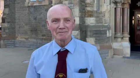 Brian is looking at the camera and is bald. He is wearing a shirt and red tie.