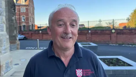 Dessie is smiling at he camera. He has a moustache and thin grey hair. He is wearing a navy polo shirt.