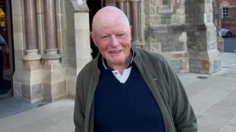 Harry is looking at the camera smiling. He is bald and wearing a green fleece and blue navy polo shirt.
