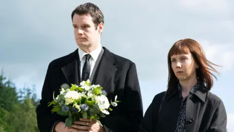 BBC/World Productions Actors Connor Swindells and Lauren Lyle in dark clothes holding flowers outside, at a memorial