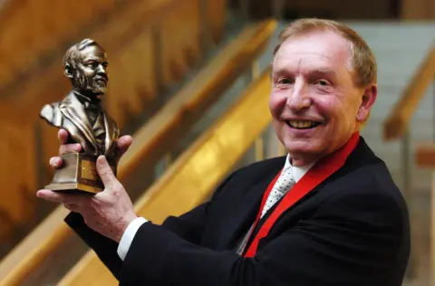 PA Media Sir Tom in dark suit and light tie holding up a bust of Andrew Carnegie. He is smiling.