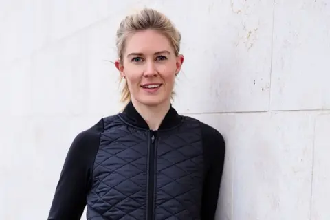 Jennifer Pybis A woman with blonde hair smiles as she leans against a white wall while wearing a black gilet.