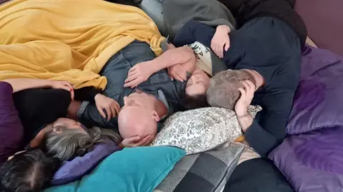 Samii Wood A group of people are covered in blankets and united in cuddles as they lie on a nest of cushions. No faces are clearly visible but there are men and women of various ages.