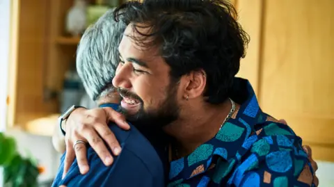 Getty Images A man with dark hair is hugging somebody else with grey hair whose face we cannot see. The man we can see is smiling.