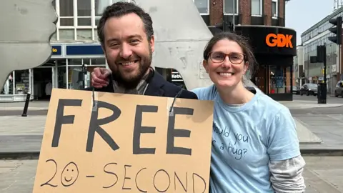 Kimberley Piper/BBC A man with short brown hair and a brown beard is smiling next to Samii who has a blue shirt and glasses with her arm around him